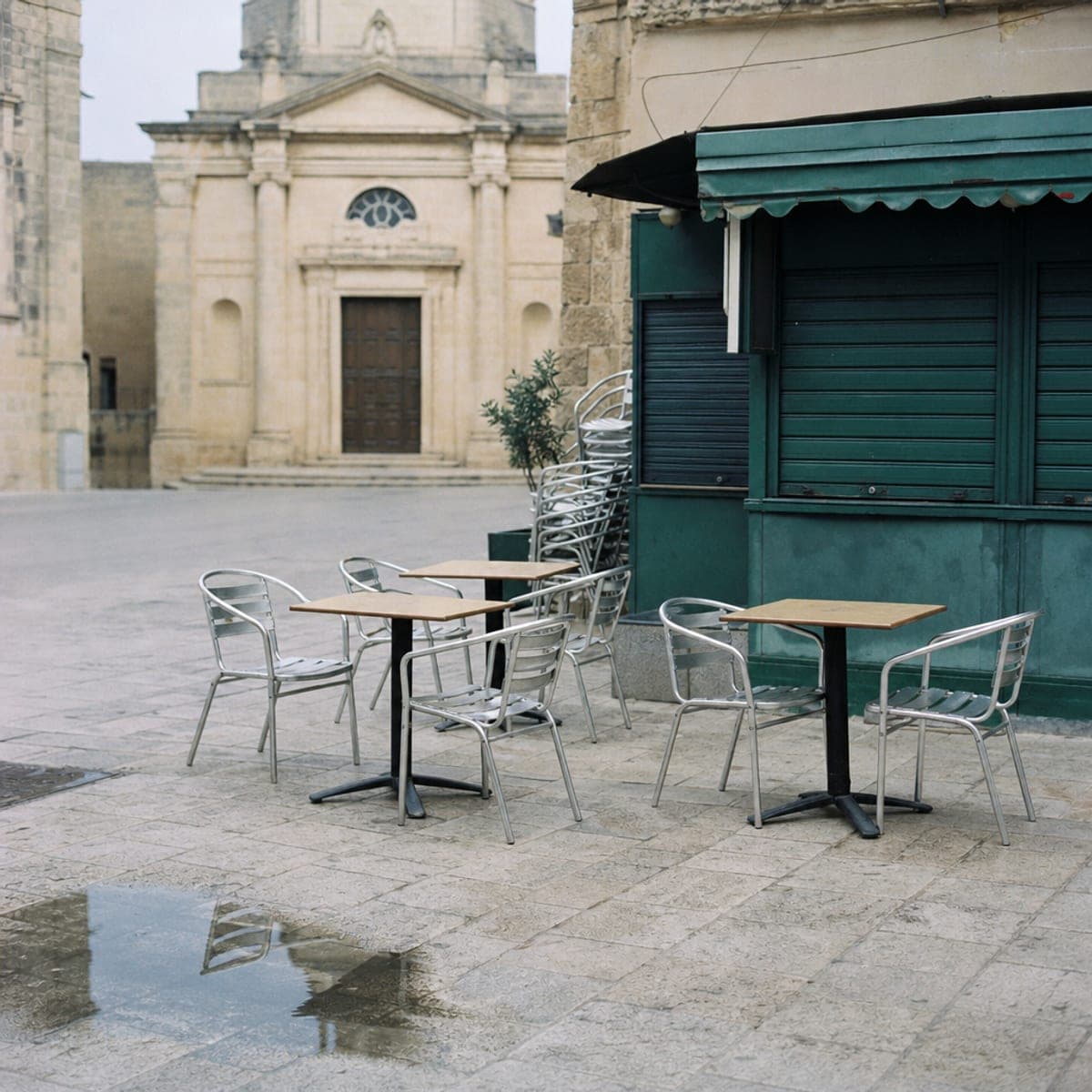 Empty café chairs on limestone paving in a Maltese village square in February, a shuttered green kiosk behind them.