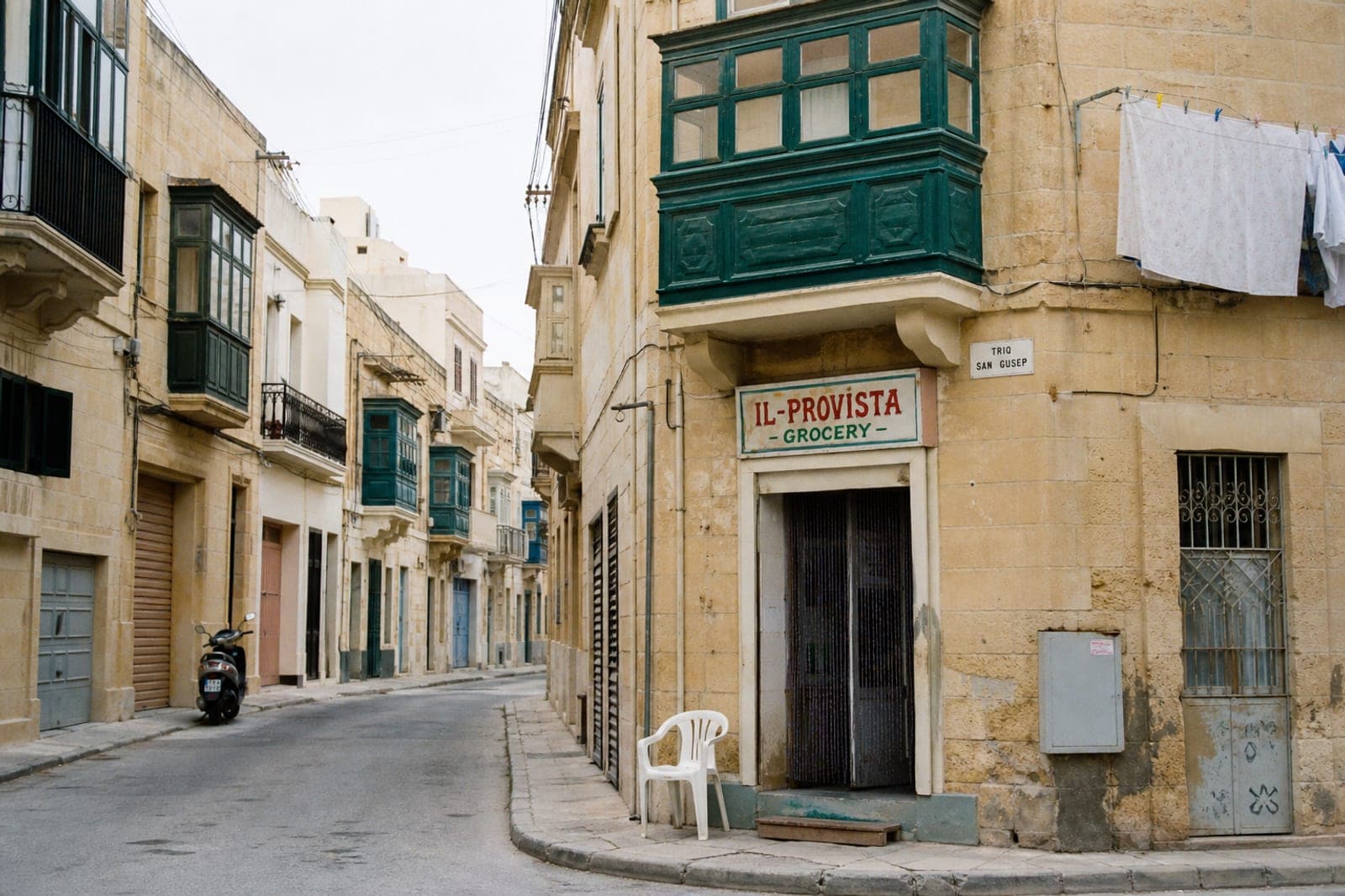 An ordinary residential street in Gżira in flat daylight: limestone walls, green wooden balconies and a small corner grocery.